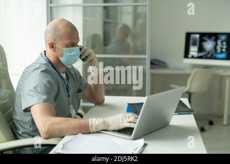 Seitenansicht des kahlen reifen männlichen Klinikers in Schutzmaske Sitzen am Schreibtisch vor dem Laptop im medizinischen Büro Und telefonieren Stockfoto