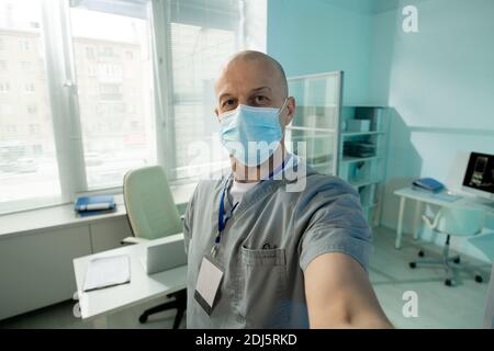 Glatze reifen männlichen Arzt in Schutzmaske Blick auf Sie Beim Selfie vor der Kamera gegen seinen Arbeitsplatz In der Arztpraxis Stockfoto