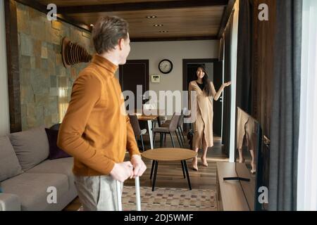 Glücklich junge asiatische Frau in eleganten beige Kleid zeigt auf Fenster, während sie ihren Mann oder Freund mit Koffer betrachten Vor der Abreise Stockfoto