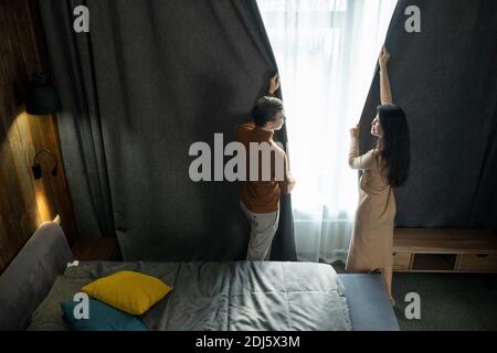 Junger Mann und seine hübsche Frau in Smart Casualwear stehen Zwischen Bett und Fenster, während Sie im Hotel dunkle Vorhänge ziehen Zimmer am Abend Stockfoto