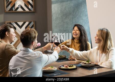 Zwei junge interkulturelle Paare in smarter Casualwear, die mit einer Brille klirren Von Rotwein über serviert Tisch zum Abendessen in luxuriös Restaurant Stockfoto