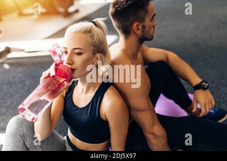 Attraktive junge Sportler halten eine Flasche Wasser, reden und lächeln, während sie sich im Fitnessstudio entspannen Stockfoto