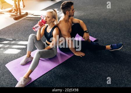 Junge Sportpaar in Sportkleidung sitzt auf Yoga-Matten und Halten Sie Wasserflaschen nach dem Training im Fitnessstudio Stockfoto