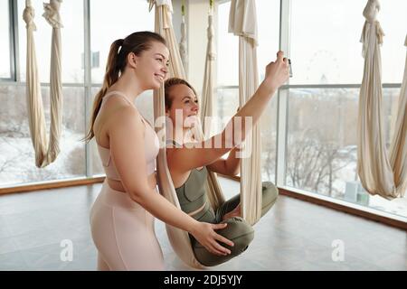 Zwei junge, fröhliche aktive Frauen in Trainingsanzügen, die das Smartphone betrachten Kamera beim Selfie im Fitnessstudio während der Yoga-Praxis fliegen Stockfoto