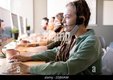 Reihe von Kundendienstmitarbeitern mit Headsets an Schreibtischen Mit Computermonitoren mit junger weiblicher Bedienerin vorne Stockfoto
