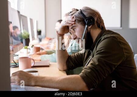 Bartbediener mit Schutzmaske unter Kinn und Headset Beratung von Kunden, während sie vor dem Computer gegen Kollegen sitzen Stockfoto