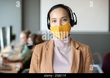 Junge weibliche Kundenbetreuer mit gemischter Rasse in Headset, Schutzmaske und Casualwear, die sich gegen eine Reihe von Kollegen stellt Stockfoto