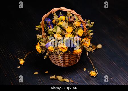 Korb mit gelben und violetten trockenen Rosen. Blumenstrauß aus verwelkelten Blumen auf einem schwarzen Holzhintergrund. Getrocknete gelbe und blaue Rosen in Herbstzusammensetzung. Stockfoto