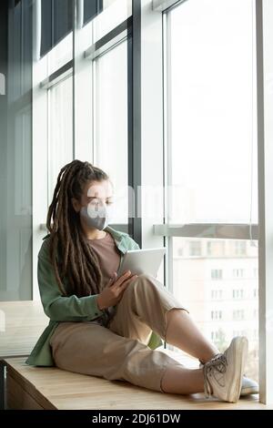 Junge Geschäftsfrau in Schutzmaske und Casualwear Blick auf Display Von digitalen Tablet beim Surfen im Netz durch große Bürofenster Stockfoto