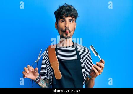 Junger hispanischer Mann trägt Friseurschürze hält Rasiermesser und Schere machen Fisch Gesicht mit Mund und schielende Augen, verrückt und komisch. Stockfoto