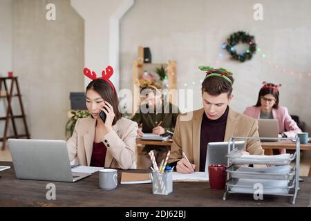 Junge zeitgenössische interkulturelle Mitarbeiter in smarter Casualwear und Weihnachtskopfbands Networking vor Laptops, während Sie in Reihen sitzen Stockfoto