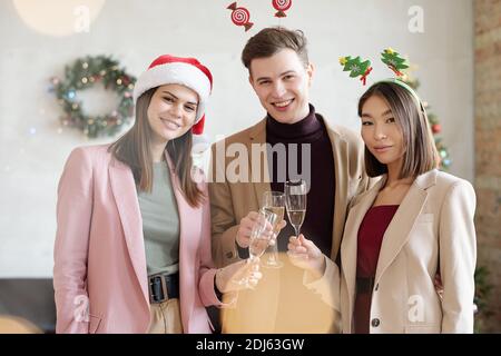 Drei junge glückliche interkulturelle Angestellte, die mit Champagner klirren Während Toast für Weihnachten und Blick auf Sie Stockfoto