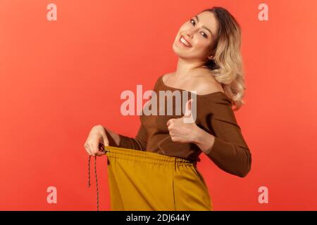 Portrait von schönen glücklich junge Erwachsene Frau haben Willenskraft zeigt schlanke Taille in großen Trousen und Daumen nach oben, erfolgreiche Gewichtsabnahme, Ernährung Konzeption. Stockfoto