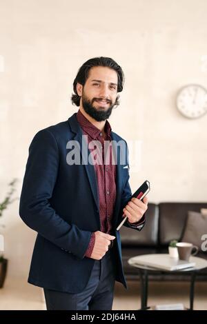 Junger erfolgreicher Geschäftsmann im eleganten blauen Anzug, der vorne steht Der Kamera im Büro gegen einen kleinen Tisch und schwarzes Leder Coach Stockfoto