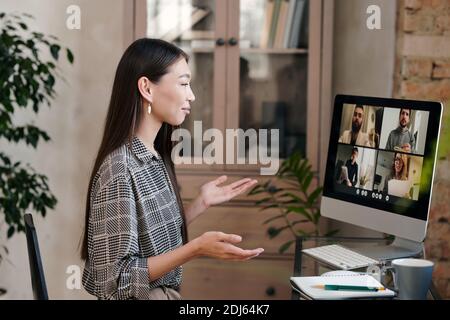 Selbstbewusst und lächelnd junge weibliche Büromanagerin mit langen dunklen Haare sitzen am Tisch und sprechen mit Kollegen am Computer Angezeigt Stockfoto