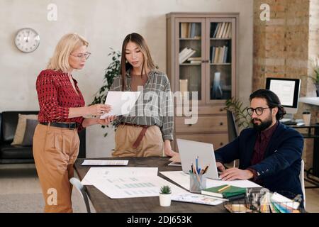 Junge und reife Weibchen stehen am Tisch und diskutieren Skizze Auf dem Papier, während bärtige Mann mit Laptop sucht online Daten Stockfoto
