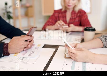 Hände von kreativen Designern oder Entwicklern mobiler Anwendungen, die sich in das Bildlauf-Programm hinein machen Smartphones beim Sitzen am Tisch und auf der Suche nach neuen Ideen Im Netz Stockfoto