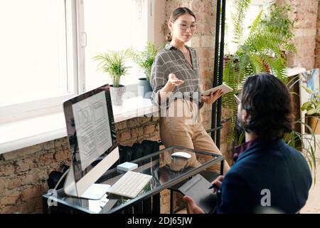 Junge weibliche mobile Anwendung Entwickler mit Tablet mit Diskussion mit Ihr männlicher Kollege zeichnet Skizze vor dem Computerbildschirm Stockfoto