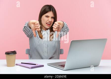 Unzufriedener Büroangestellter zeigt Daumen nach unten, mag Geste nicht, äußert Missbilligung, kritisiert sitzen am Arbeitsplatz mit Laptop. Stehbolzen für den Innenbereich Stockfoto