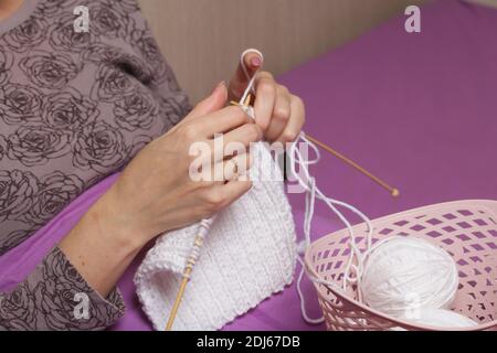 Die Frau strickt. In meinem Schlafanzug unter den Bezügen sitzend. In der Nähe befindet sich ein Korb mit Fäden. Nahaufnahme. Stockfoto