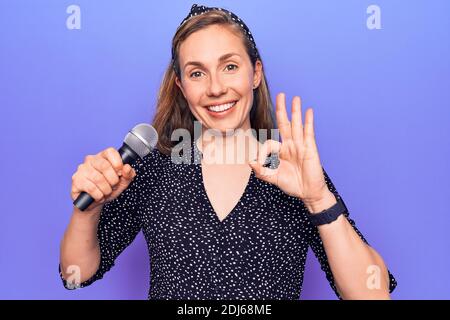 Junge schöne blonde Frau hält Mikrofon singen über lila Hintergrund tun ok Zeichen mit Fingern, lächelnd freundlich gestikulieren ausgezeichnete Symbol Stockfoto