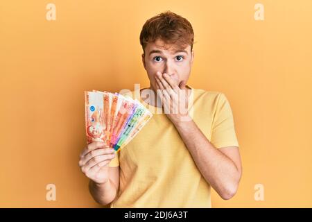 Junger kaukasischer Mann hält philippinische Peso-Banknoten, die den Mund mit der Hand bedecken, schockiert und Angst vor Fehlern. Überraschter Ausdruck Stockfoto