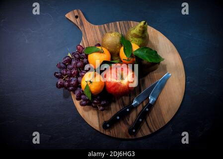 Sammlung von Obst auf einem Nussbaum Holz Schneidebrett mit Schneidmesser. Frucht enthält Apfel, Mandarine, Trauben und Birne. Stockfoto