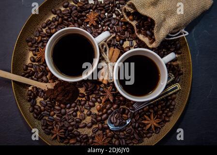 Draufsicht auf zwei Tassen Kaffee auf einem goldenen Serviertablett, umgeben von geröstetem Kaffee, gemahlenem Kaffee, Sackleinen mit gerösteten Bohnen und Gewürzen. Stockfoto