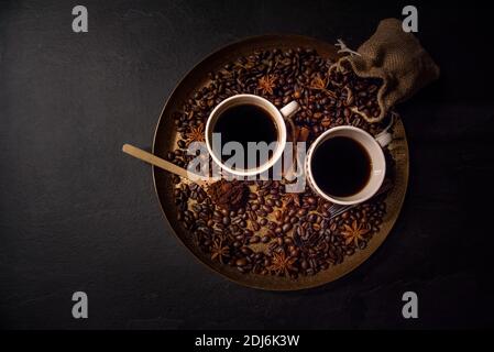 Draufsicht auf zwei Tassen Kaffee auf einem goldenen Serviertablett, umgeben von geröstetem Kaffee, gemahlenem Kaffee, Sackleinen mit gerösteten Bohnen und Gewürzen. Stockfoto
