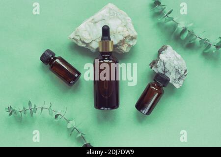 Schwarze Flaschen aus Glas ohne Marken mit kosmetischem Öl und ätherischen Ölen auf Natursteinen, grüner Hintergrund mit Eukalyptusblättern, Draufsicht. Aromatherapie Stockfoto