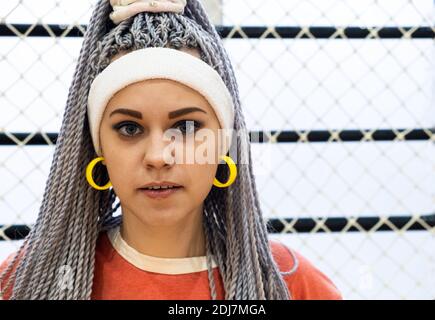 Porträt von jungen stilvollen Frau mit canecalon Angst Zöpfe und Gelbe Ohrringe blicken mit einer neutralen Gesichtsfarbe in die Kamera Ausdruck Stockfoto