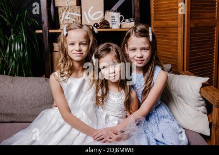 Zwei niedliche Schwestern in weißen Kleidern sitzen auf dem Sofa dicht beieinander und spielen zu Hause. Kleine Schwester umarmt ihre kleinste Schwester. Stockfoto