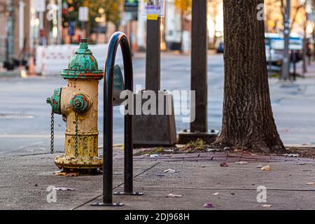 Ein rostiger, alter Hydrant an der Seite einer Straße auf dem Bürgersteig mit abgeplatteten gelben und grünen Farben darauf. Im unscharfen Hintergrund ist eine Staue Stockfoto