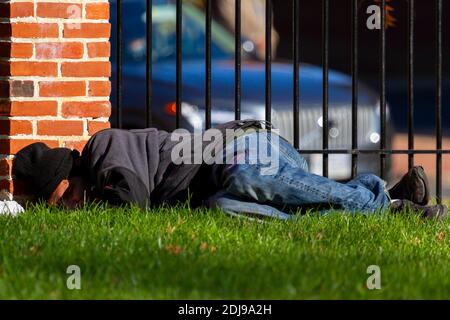 Alexandria, VA, USA 11-28-2020: Ein obdachloser Latino-Mann mit Sweatshirt und Jeans schläft auf dem Gras im Freien in einem Stadtpark an einer Ziegelmauer A Stockfoto