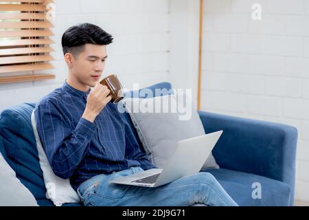 Junge asiatische Geschäftsmann lächeln und arbeiten von zu Hause aus mit Laptop-Computer online auf Sofa im Wohnzimmer, freiberuflicher Geschäftsmann mit Notebook und trinken c Stockfoto