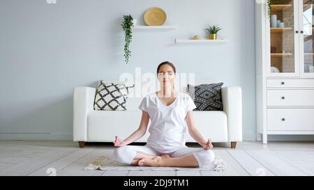 Ruhige junge schwangere Frau in weiß meditiert im Lotus sitzen Pose auf dem Teppich neben Designer-Sofa in geräumigen Zimmer Zu Hause Stockfoto