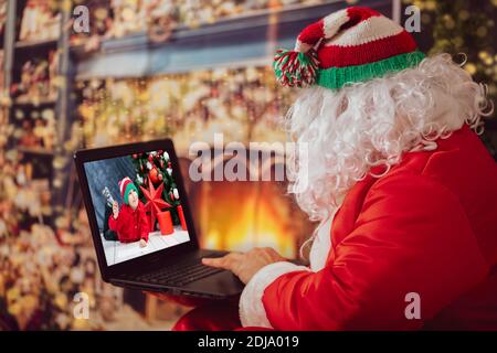 Der Weihnachtsmann schaut auf den Laptop. Ein Mann in einem Weihnachtsanzug am Computer auf dem Hintergrund der Neujahrsbeleuchtung. Der moderne Zauberer. Weihnachten h Stockfoto