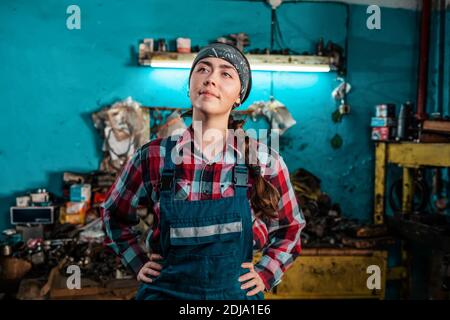 Porträt einer jungen schönen Mechanikerin in Uniform, die mit den Händen auf den Hüften posiert und lächelt. Arbeitszimmer im Hintergrund. Das Konzept Stockfoto
