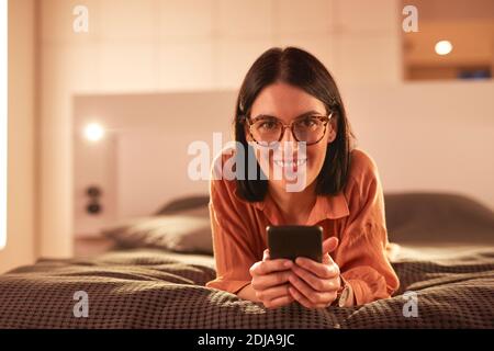 Porträt einer zeitgenössischen erwachsenen Frau mit Smartphone, während auf dem Bett in minimalem Innenraum und lächelnd auf Kamera liegen, kopieren Raum Stockfoto