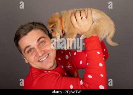 Junge gut aussehende Mann mit Weihnachten Stil Outfit hält eine nette Kleiner Welpe auf grauem Hintergrund Stockfoto