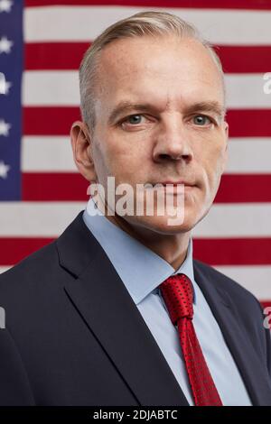 : Vertikales Porträt eines reifen Politikers, der vor dem Hintergrund der US-Flagge die Kamera anschaut Stockfoto