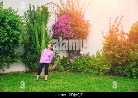 Frau steht am Baum mit violetten Blumen. Die Frau auf dem Rasen neben dem Kaktus. Stockfoto