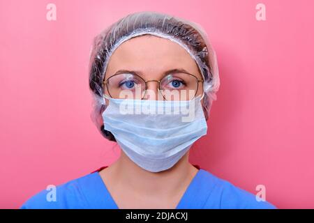 Porträt einer Ärztin in Brille und einem medizinischen Hut, Nahaufnahme. Krankenschwester in blauer Uniform und Schutzmaske auf rosa Hintergrund. Stockfoto