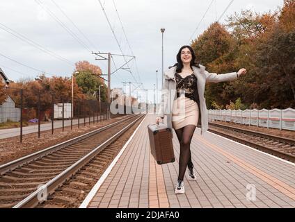 Junge lächelnde Mädchen in einem grauen Mantel mit einem Koffer Spaziergang am Herbsttag entlang des Bahnhofs Stockfoto