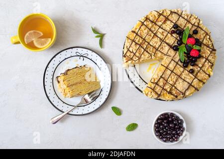 Mehrschichtiger Vanillekuchen mit Beeren, Minze verziert. Ein Stück leckeren Kuchen auf einem Teller und eine Tasse Morgentee mit Zitrone. Draufsicht. Stockfoto