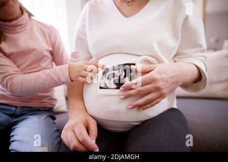 Nahaufnahme der schwangeren Mutter und Tochter, die das Ultraschallbild des Babys auf dem Bauch halten. Stockfoto