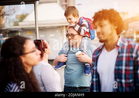 Der junge, gutaussehende Vater hält sein kleines, entzückendes Kleinkind auf seinen Schultern und hält eine Tasse Kaffee, während er mit einer Frau auf einer Straße spricht. Stockfoto