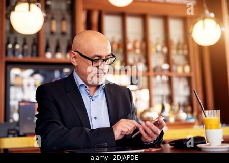 Reifer professioneller seriöser Geschäftsmann benutzt ein Telefon und trinkt Kaffee und Saft, während er im Coffee Shop sitzt. Stockfoto
