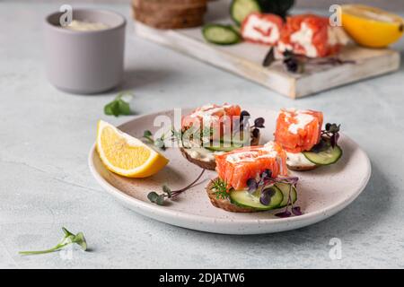 Mini-Sandwiches mit geräuchertem Lachs, Frischkäse, Gurke und Microgreen auf Roggenbrot, grauer Steinhintergrund. Vorspeise mit Lachs und Käse c Stockfoto