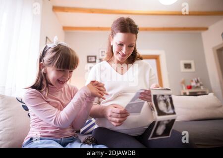 Schöne schwangere Mutter und kleine entzückende Tochter Blick aufgeregt auf die Ultraschallbilder des Babys, während sie auf dem Sofa zu Hause sitzen. Stockfoto